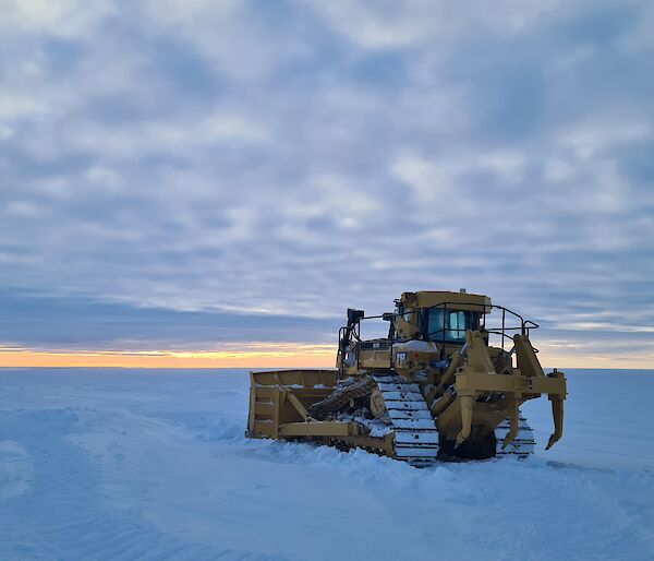 A large yellow bulldozer parked on a flat expanse of snow covered ice. There is an even layer of low cloud stretching out to the horizon.