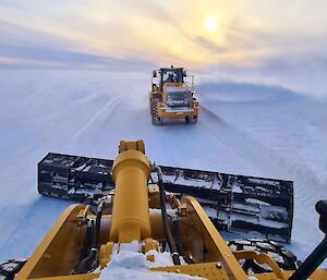 Photo taken from the driver's seat of a large grading machine. The machine has a blade on the front which clears snow from the runway, and levels the ground. In front of this is another machine that is helping clear the snow.