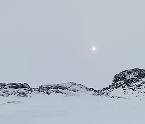 A rocky, snow and ice covered island is visible rising from a frozen sea. The sky is cloudy and a muted white colour with the sun visible as a white dot through the clouds