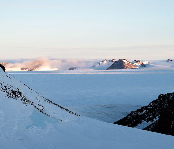 A maintain range with snow being blown across it is visible in the distance eover an ice plateau. In the foreground are more rocky mountains.