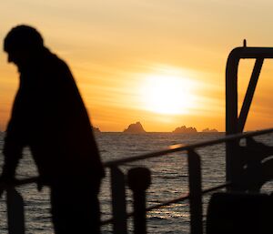 A man is standing against a ship's railing looking at the sea. In the distance the sun is low on the horizon and there are icebergs silhouetted against the sunset