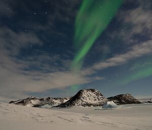 A rocky, hilly, island rises from a frozen sea. In the sky a bright green aurora is visible against the stars and clouds