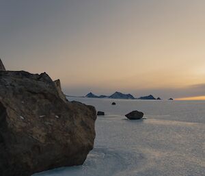 Some rocks are in the foreground with a vast ice plateau stretching to the horizon. A rocky mountain range is seen in the far distance and the glow from a sunset highlights the horizon.