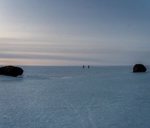 Two people are in the distance on a vast ice plateau. In the foreground to the left and right of frame are two large rocks.