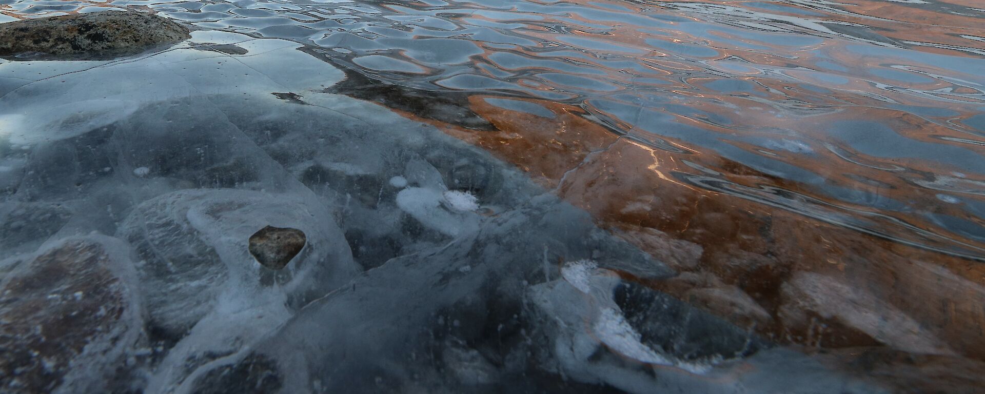 Frozen lake water highlights rocks in a frozen lake. Colours reflect upon the frozen surface.