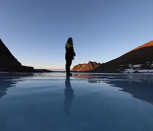 A woman is standing on a highly reflective, frozen lake. There are rocky hills  in the background to the left and right of frame
