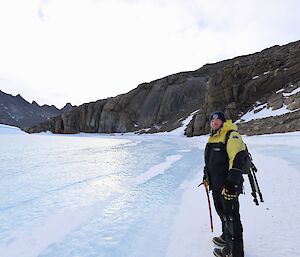 A man is standing on a frozen lake. In the background is a rocky landscape rising from the ice.