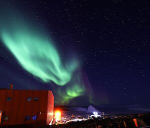 A bright green and puple aurora is visible in the sky above a large red building. In the distance two icebergs are visible locked into a frozen sea