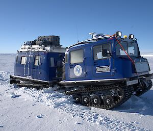 A blue Hägglunds vehicle is struggling to drive through a deep snow drift on the sea ice