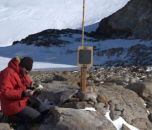 A man is kneeling next to a proclamation marker and reading a rolled piece of paper. In the background a glacier rises against a rocky landscape