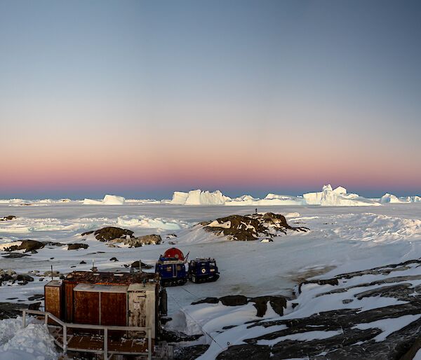 A wide panorama photo with a rocky, snow and ice covered island in the foreground with a hut in the middle of frame and a blue Hägglunds vehicle parked behind it. In the distance are a large number of icebergs frozen into the sea ice.