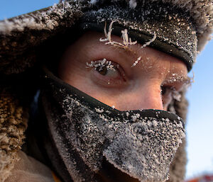 A lose up of a man looking at the camera. He is wearing a furry hat and face mask across his lower face. His eyelashes and hair are frozen and the mask is covered in snow.