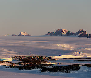 In the distance a multicoloured station can be seen on a rocky landscape. Behind it, an ice plateau rises with jagged, rocky, mountain ranges in the far distance.