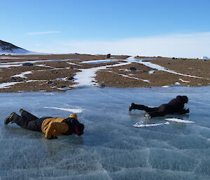 Two men are laying on a frozen lake. One appears to be licking the ice. In the background is a rocky island.