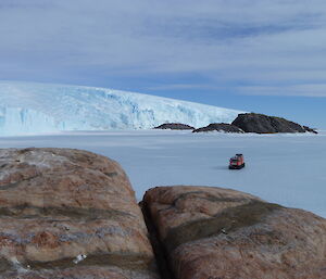 On the sea ice is parked a bright orange Hägglunds vehicle. In the distance is a rocky island and a large glacier rising above the ice.