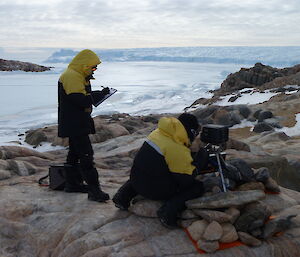 Two men are working on an automated camera that is mounted on a tripod on a rocky island. In the distance is a large glacier