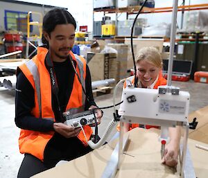 A man and woman in orange work vests looking at a scientific instrument in a warehouse.