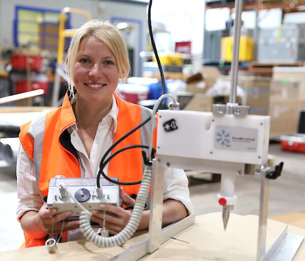 A woman in an orange work vest holding a small control box for a snow measurement instrument.