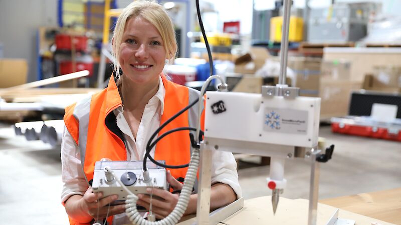 A woman in an orange work vest holding a small control box for a snow measurement instrument.