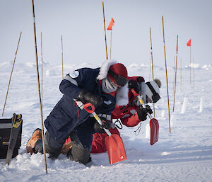 Two people kneeling in snow and holding shovels.