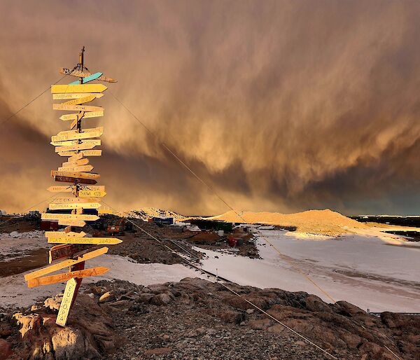 A post covered from top to bottom in town names and distances stands on top of a rocky hill. In the background the sky is filled with a large stormy cloud and the ground is covered in snow.