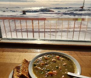 A bowl of soup and some toast sitting on a bench in front of a window. The view out the window is snow covered ground and blue sky. In the distance are low icebergs.