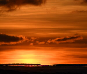 The sun has just set and the sky is lit up with a range of orange, yellow and red. There are two long low icebergs silhouetted against the sky.