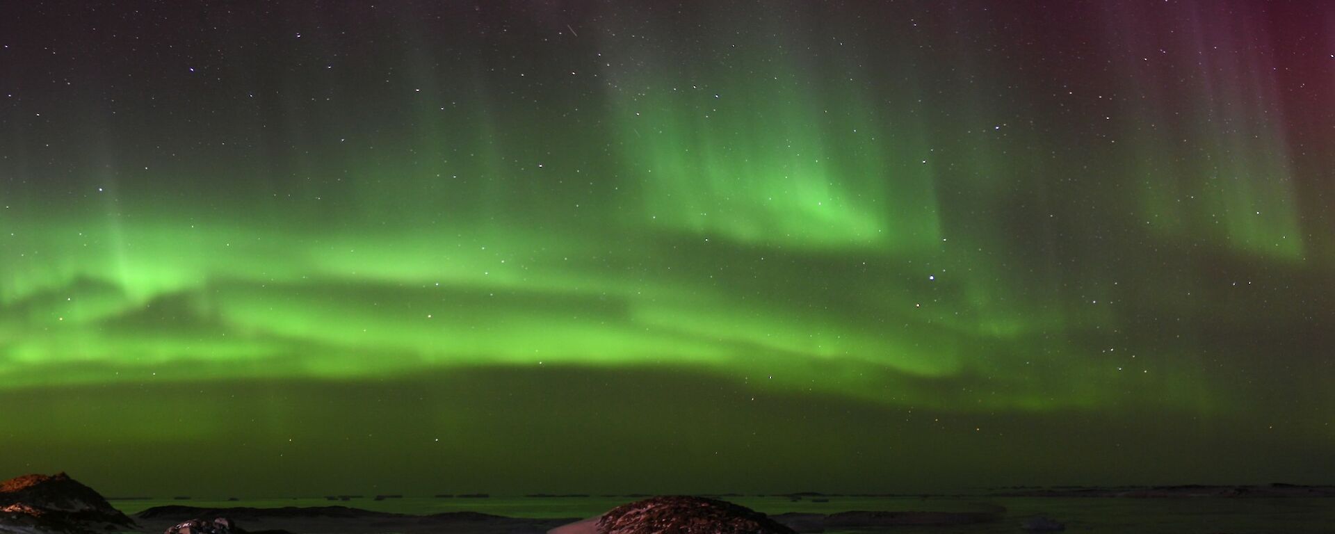 A vivid green band of aurora lights the sky in this panoramic photo. To the left of the photo is a collection of buildings that is Casey Station.