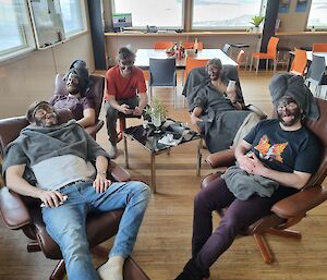 a group of five people sitting on chairs with brown coloured coffee facials on their faces and some with towels on their heads.