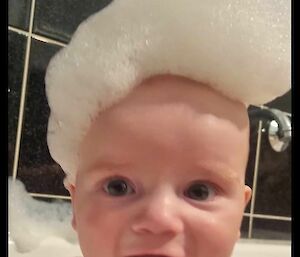 A smiling baby with a mass of soapy bubbles on top of his head