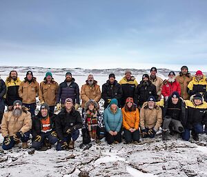 Two rows of people, back row standing, front row crouched down, stand in their winter clothes in a winter setting of snow, a frozen bay and snow-covered hills in the background.