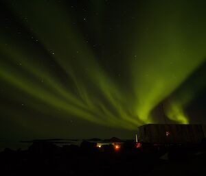 A bright green aurora is visible in the night sky above an older building which has a red glow in the window