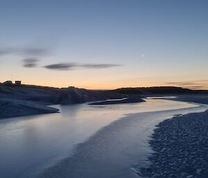 A sunset reflects upon water that is lying in a crack between the sea ice and the rocky land to the left of frame.