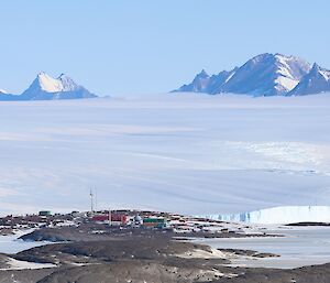 A coloured station with a number of brightly coloured buildings and a large wind turbine is visible in the distance on a rocky landscape surrounded by ice. In the far distance a number of rocky mountains can be seen rising from the ice plateau.