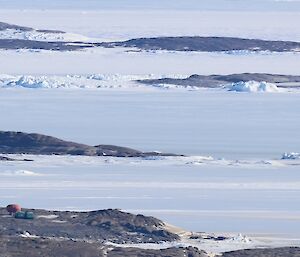 A number of small oval shaped huts and a green Hägglunds vehicle can be seen in the distance on a rocky island. The ocean is frozen over with sea ice.
