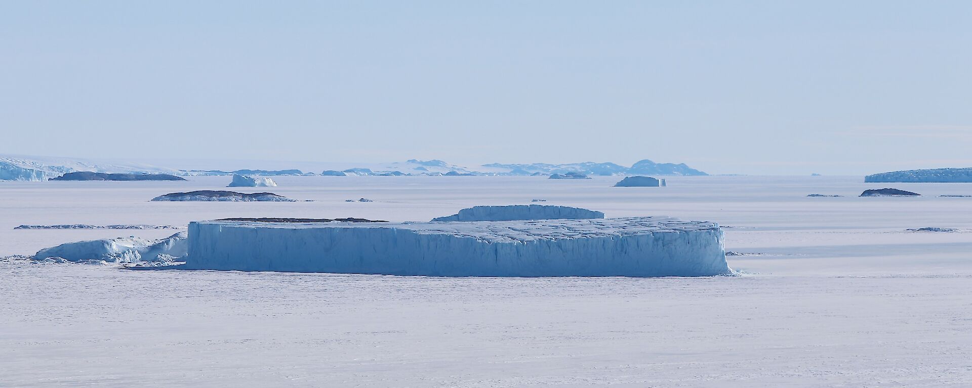 A large iceberg can be seen frozen into the sea-ice that surrounds it. In the distance are other icebergs and three small rocky islands.