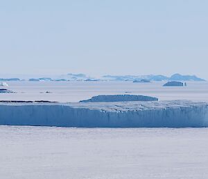 A large iceberg can be seen frozen into the sea-ice that surrounds it. In the distance are other icebergs and three small rocky islands.