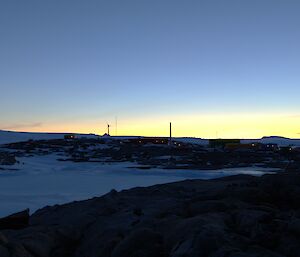 The sunset in the distance highlights a number of station buildings on a rocky landscape. A large, three bladed, wind turbine is visible against the horizon.