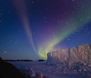 A bright green and purple aurora is visible in the night sky above an iceberg that is frozen into the sea ice. The glow of sunrise is brightening the night sky.