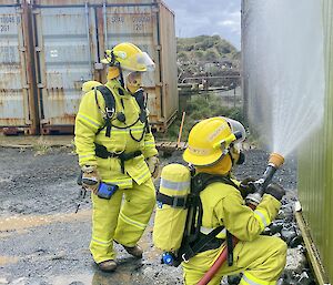 Two people in yellow firefighting gear, aim a hose at a building