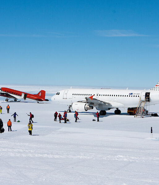 A white airplane on the ice runway with a red small plane in the distance and passengers walking off