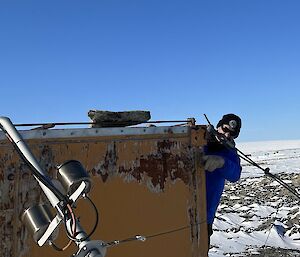 A man is standing on the side of a rustic hut, looking at a broken mast.