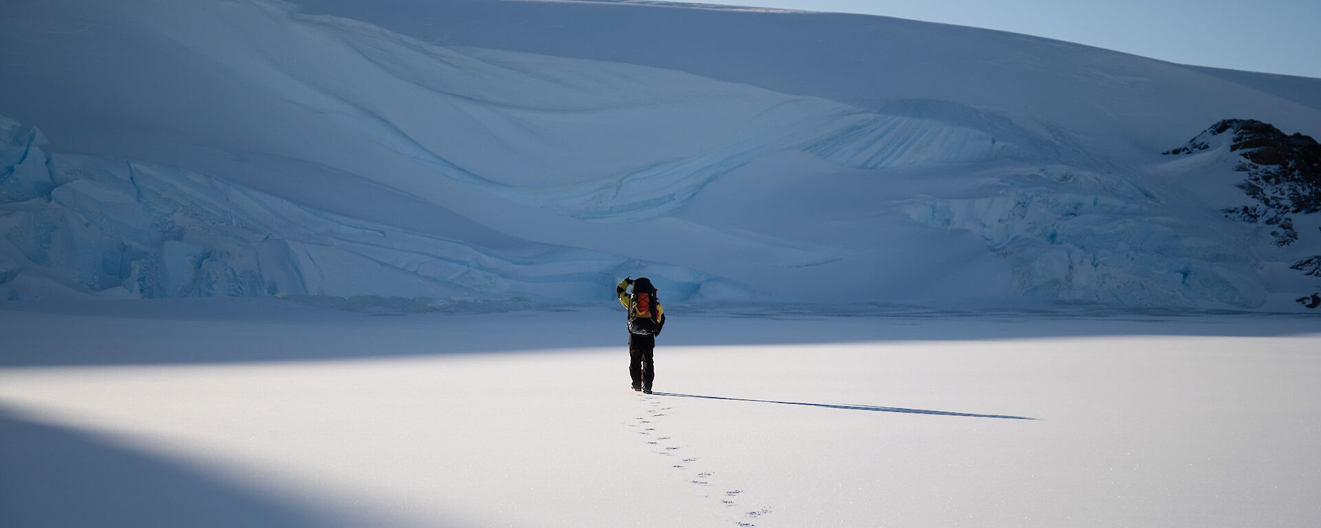 A behind view of an Antarctic expeditioner walking in the snow towards a large snowy ridge