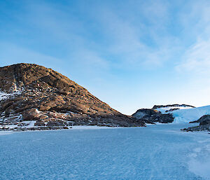 A frozen blue lake in the foreground with a large rocky outcrop in the background and clear blue skies above.