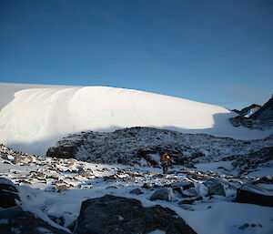 A large accumulation of snow off the edge of a ridge looks like a curling wave. Snow covered rocks are visible in the foreground.