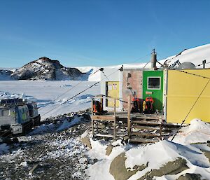 A green, yellow and grey field hut sits in a rocky and snow-covered landscape. A Hagglunds vehicle is parked to the left of it.