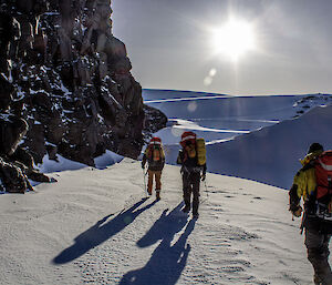 A behind view of three Antarctic expeditioners walking off into snow covered hills with a large rock wall to their left