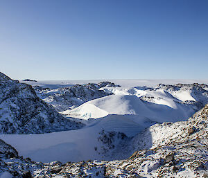 A snow-covered ridge surrounded by rocky snow-covered hills with a large ice plateau in the background.