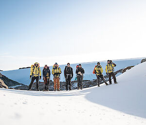 Seven people wearing snow gear standing in a row in a snow covered landscape.