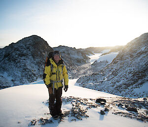 A smiling man stands atop a snow covered peak with a glacial valley in the background.